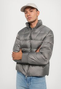 Jachetă puf gri cu guler înalt, închidere cu fermoar și manșete ribalte. Are un design elegant cu detalii subtile de model geometric.