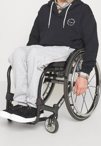 Felpa nera con cordini bianchi, pantaloni grigi e sneakers nere. Individuo seduto su una sedia a rotelle con una mano sulla ruota.