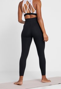 Leggings neri a vita alta e un reggiseno sportivo bianco con design a incrocio sulla schiena, indossati da una persona in piedi a piedi nudi su un tappetino da yoga.