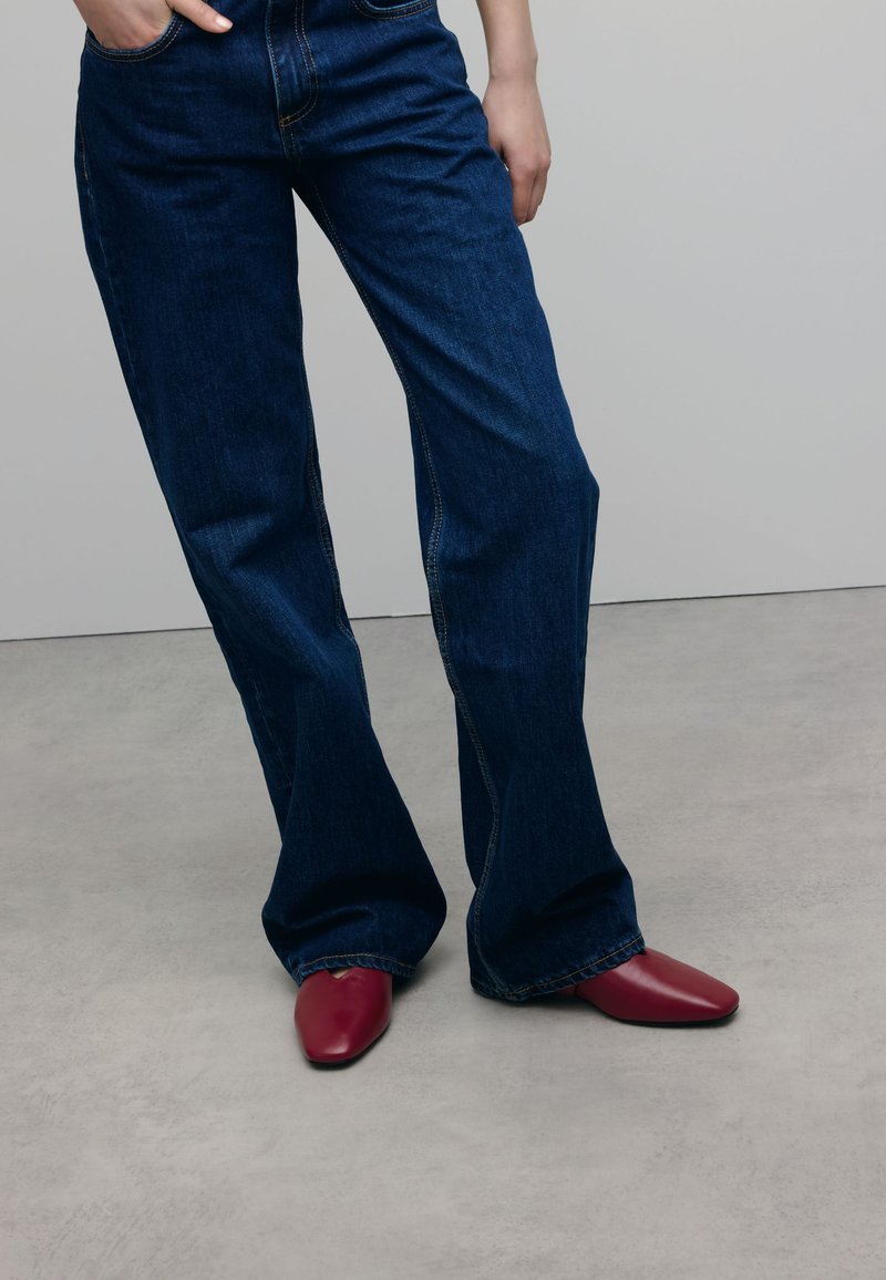 Donkerblauwe spijkerbroek met een flared pasvorm en contrasterende oranje stiksels, gecombineerd met bordeauxleren schoenen met een puntige neus op een lichtgrijze ondergrond.