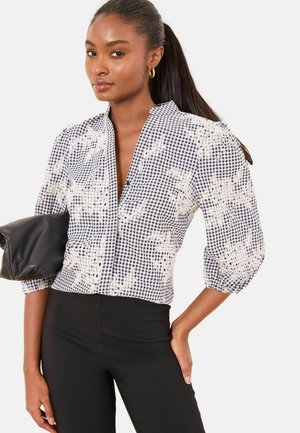 Sort og hvid gingham-bluse med puffede ærmer, blomsterbroderi og dyb V-hals. Model holder en sort clutch-taske og har sorte, figursyede bukser på.