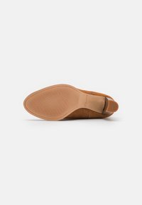 Zapato marrón de ante con punta afilada, suela de goma texturizada y tacón bajo, mostrando la marca "Clarks" en la suela sobre un fondo blanco.
