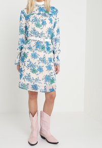 Robe fleurie avec des motifs bleus et verts sur un fond clair ; manches longues ; taille cintrée avec une ceinture blanche ; associée à des bottes de cowboy roses.