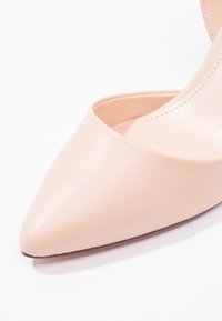 Chaussure à talon haut rose pâle à bout pointu, avec une texture en cuir lisse et une découpe latérale, sur un fond blanc uni.
