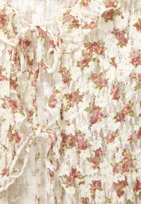 Top in pizzo con stampa floreale in rosa e verde su sfondo crema. Presenta lacci all'altezza dello scollo e bordi arricciati. Texture intricata.