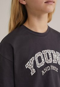 Tamsiai pilka medvilninė marškinėliai su trumpomis rankovėmis, kuriuose užrašyta "YOUNG" leopardų raštu ir "AND FREE" balta siuvinėjimu. Apvalus kaklas, laisvas siluetas.