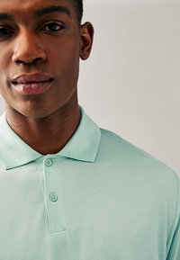 Ljusgrön pikétröja med ett mönstrat tyg, med krage och två knappar vid halsen. Närbild som visar tygdetaljerna.