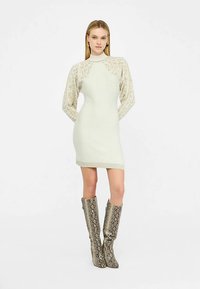 Stickad klänning i ljusgrönt med mönstrade, transparenta ärmar, hög halsringning och figursydd form. Par knähöga, glänsande stövlar med ormskinsmönster.