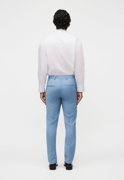 Άνδρας με σκούρα σγουρά μαλλιά, που φοράει λευκό πουκάμισο με μακριά μανίκια, κρυμμένο μέσα σε ανοιχτόχρωμο μπλε παντελόνι και καφέ επίσημα παπούτσια, στέκεται γυρισμένος προς τα πίσω.