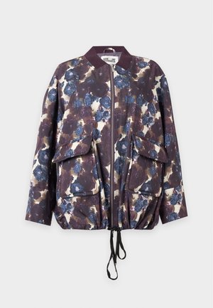 Persoon draagt een donker gebloemde oversized bomberjack, witte broek met wijd uitlopende pijpen en puntige bruine schoenen, staand met één hand in de zak van het jack.