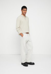 Tricou polo cu mânecă lungă, de culoare bej deschis, cu guler, asortat cu pantaloni largi de culoare gri deschis și pantofi negri. Design simplu, cu detalii minimale.