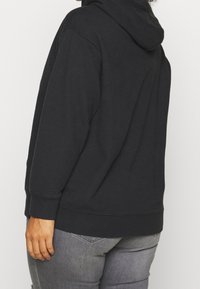 Persona che indossa un cappuccio nero e jeans grigi, vista di spalle contro uno sfondo chiaro e uniforme.