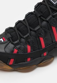 Baskets noires avec un dessus en cuir texturé, des bandes accentuées rouges, une doublure en mesh noir et une semelle en caoutchouc avec une section transparente.