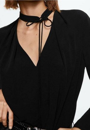 Frau trägt ein schwarzes V-Ausschnitt-Oberteil mit einem gebundenen schwarzen Choker und einer schwarzen Jacke, die Hand ruht auf einem mit Nieten besetzten schwarzen Gürtel.