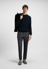 Navy textured sweater, gray tailored trousers with sharp creases, and gray slip-on shoes; model holds a black jacket over the shoulder.