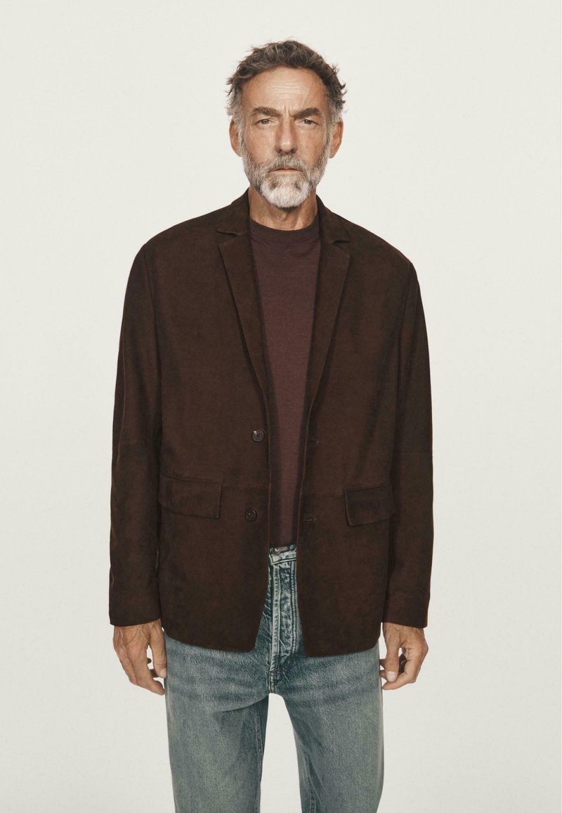 Blazer in suede marrone con due tasche frontali, chiusura a un bottone e rever a punta, indossato sopra una camicia bordeaux e abbinato a jeans azzurri chiari.