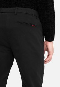 Person wearing black pants with a back pocket featuring a small red fabric detail and a textured black long-sleeve top.