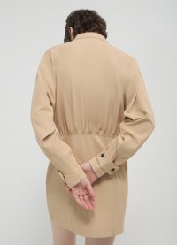 Beige leichte Jacke mit tailliertem Schnitt, langen Ärmeln und Knöpfen an den Bündchen. Glatte Textur mit Kragen und mittellanger Länge bis zur Oberschenkellinie.