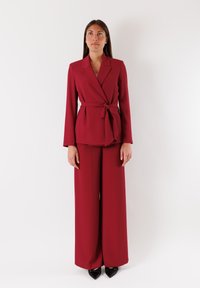 Blazer sartorial borgogna con cintura annodata in vita, abbinato a pantaloni a gamba larga. Tessuto liscio; maniche lunghe; tacchi neri a punta.