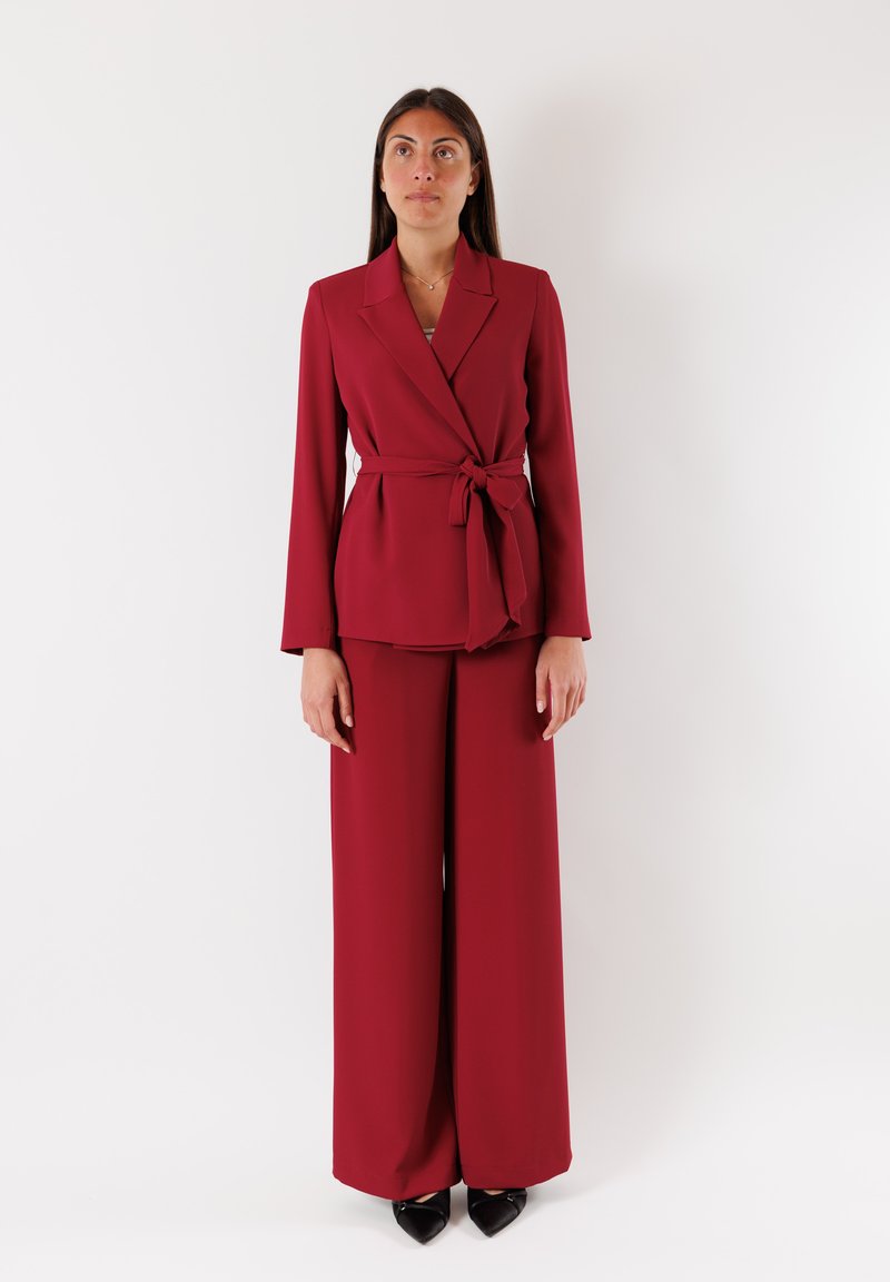 Blazer sartorial borgogna con cintura annodata in vita, abbinato a pantaloni a gamba larga. Tessuto liscio; maniche lunghe; tacchi neri a punta.