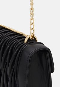 Černá kožená kabelka s plisovaným designem, zdobená zlatým řetízkem a kulatým kovovým detailem. Hladká textura a pevný tvar.