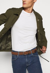 Chaqueta verde oliva, camiseta blanca, jeans de mezclilla azules y un cinturón de mimbre marrón. La chaqueta tiene botones a presión y un interior suave y texturizado.