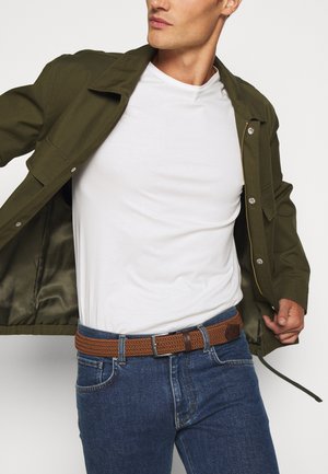 Olivgrüne Jacke, weißes T-Shirt, blaue Jeans und ein brauner geflochtener Gürtel. Die Jacke hat Druckknöpfe und ein glattes, strukturiertes Innenfutter.