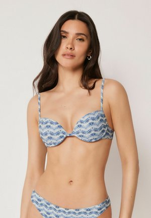 Kobieta z długimi ciemnymi włosami, nosząca niebieski i biały wzorzysty, wyściełany top bikini i dopasowane doły na jednolitym tle.