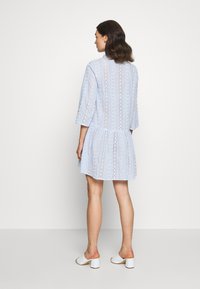 Vestido azul claro com design de ilhós, saia em camadas e mangas 3/4. Textura suave, padrão subtil e bainha arredondada. Mule brancas de salto.