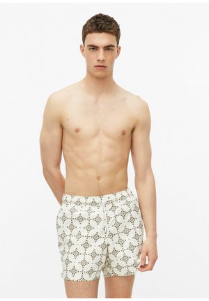 Jeune homme aux cheveux courts et bouclés portant un short de bain blanc avec un motif géométrique marron, debout devant un fond blanc uni.