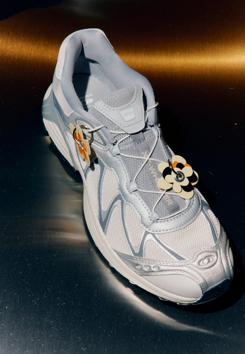 Lys grå treningssko med meshstoff, sølvdetaljer og mønstret såle. Har en dekorativ blomster-tag på skolissene.