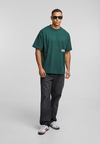 Grønn bomull t-skjorte med løs passform, korte ermer, og en liten hvit logo. Matchet med mørke denimbukser og grå sneakers.