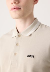 Beige Poloshirt aus strukturiertem Stoff, mit einem Zwei-Knopf-Verschluss, kontrastierendem cremefarbenem Kragen und schwarzem Stickerei des Brands "BOSS."