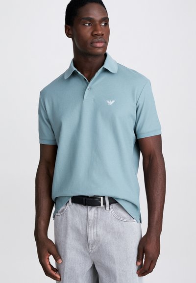 Polo di colore azzurro chiaro con colletto a punta, maniche corte e un logo discreto sul petto, abbinato a jeans grigi e una cintura nera.