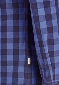 Camisa a cuadros azules con una mezcla de cuadros en tonos de azul claro y oscuro. Incluye una etiqueta en el costado con el nombre de la marca. Textura de tela suave.