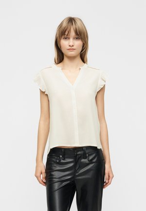 Młoda kobieta ubrana w białą bluzkę z krótkim rękawem i falbankami na ramionach oraz czarne skórzane spodnie, stojąca na jednolitym jasnym tle.