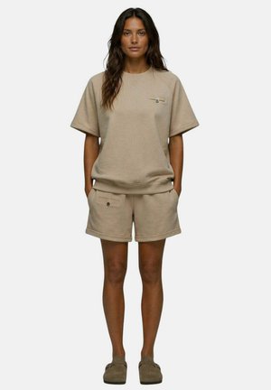 Femme debout, mains dans les poches, portant un sweat-shirt beige à manches courtes, un short assorti avec poches boutonnées et des chaussures faciles à enfiler.