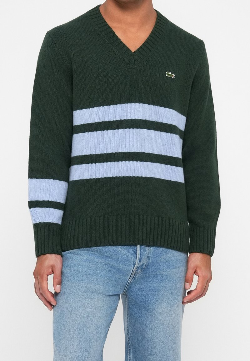 Personne portant un pull vert foncé à col en V avec des rayures bleu clair et vert clair sur la poitrine et la manche gauche, associé à un jean bleu clair.