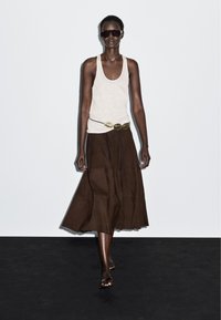 Lange persoon met donkere zonnebril, wit tanktop, bruine midirok met gouden gesp riem en sandalen met transparante band loopt over een zwarte vloer.