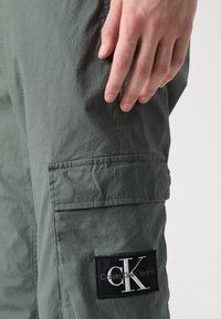 Spodnie typu cargo z przygaszonego zielonego materiału, z boczną kieszenią z czarną naszywką z logo "CK" oraz teksturowaną powierzchnią.