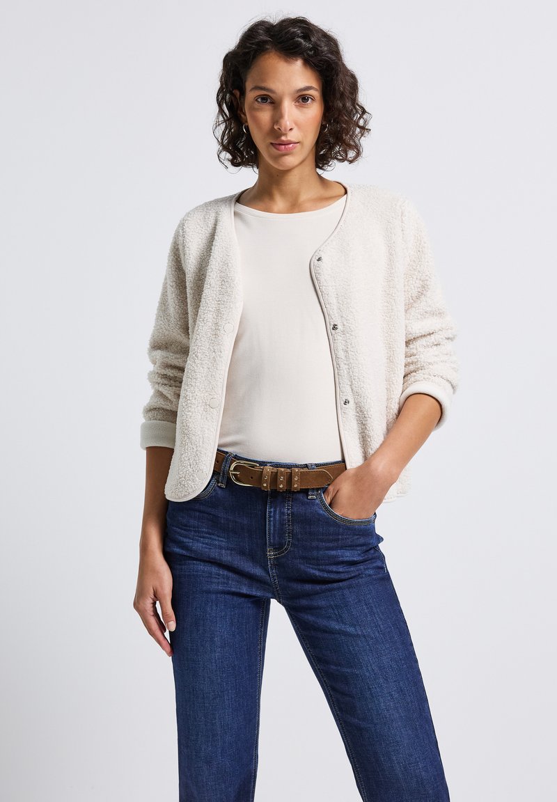 Cárdigan texturizado color crema con botones, combinado con una blusa clara y jeans oscuros. Cuenta con un cinturón marrón que enfatiza un ajuste casual.