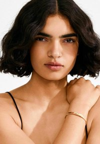 Kuldne metallist käevõru tekstuurse, haamritud viimistlusega, kantud keskmise nahatooniga ja laineliste tumedate juustega inimese randmel.