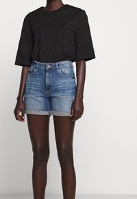 Mörkhyad person som bär en svart kortärmad skjorta och höga, uppvikta jeansshorts mot en enfärgad ljus bakgrund.