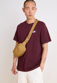 Ruskea olkalaukku säädettävällä hihnalla, teksturoidsella kankaalla ja etutaskulla. Mallilla on päällään marjapunaista t-paitaa, jossa on valkoinen Nike-logo, ja vaaleat shortsit.
