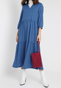 Modré vzorované šaty s tříčtvrtečními rukávy, výstřihem do V, nařaseným designem, doplněné burgundskou kabelkou a bílými botami na blokovém podpatku.