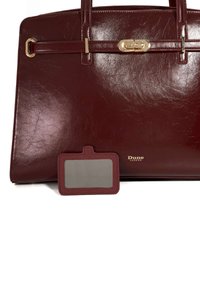 Burgundy leather handbag with a structured design, gold hardware accents, and a matching luggage tag. Smooth texture and polished finish.