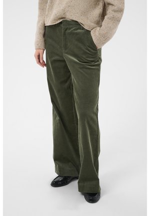 Personne portant un pantalon large en velours côtelé vert, un pull en maille beige et des chaussures plates noires, debout avec une main dans la poche.