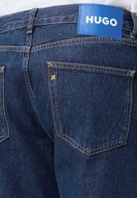 Σκούρο μπλε τζιν denim, πίσω όψη με κίτρινο ράψιμο και μπλε δερμάτινο ένθετο με την ετικέτα "HUGO" στη μέση.