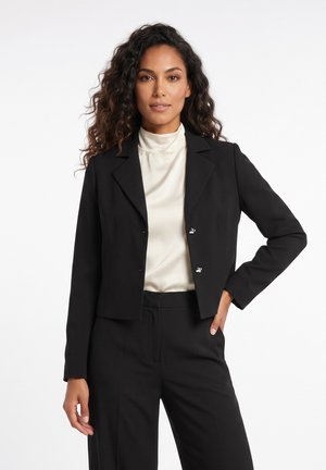 Femme aux cheveux bouclés portant un blazer noir court sur un chemisier blanc à col haut et un pantalon noir, debout avec une main dans la poche.