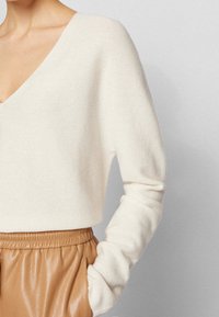 Pull à col en V de couleur crème en matière tricotée, doté de manches longues et d'une coupe décontractée, associé à un pantalon en cuir marron clair.